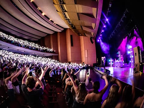 Lied Center interior with vibrant lighting, cheering crowd with phones raised, and performers on stage.