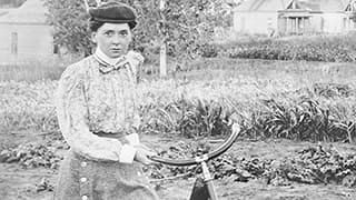Louise Pound in vintage attire holding a bicycle in a rural setting with houses and vegetation in the background.