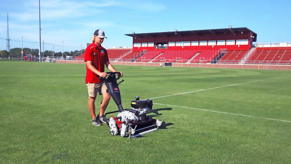 A young man mows the field at a soccer stadium. He is wearing a red Nebraska polo with a white "N" on it, brown shorts, black shoes and a black-and-gray baseball cap.