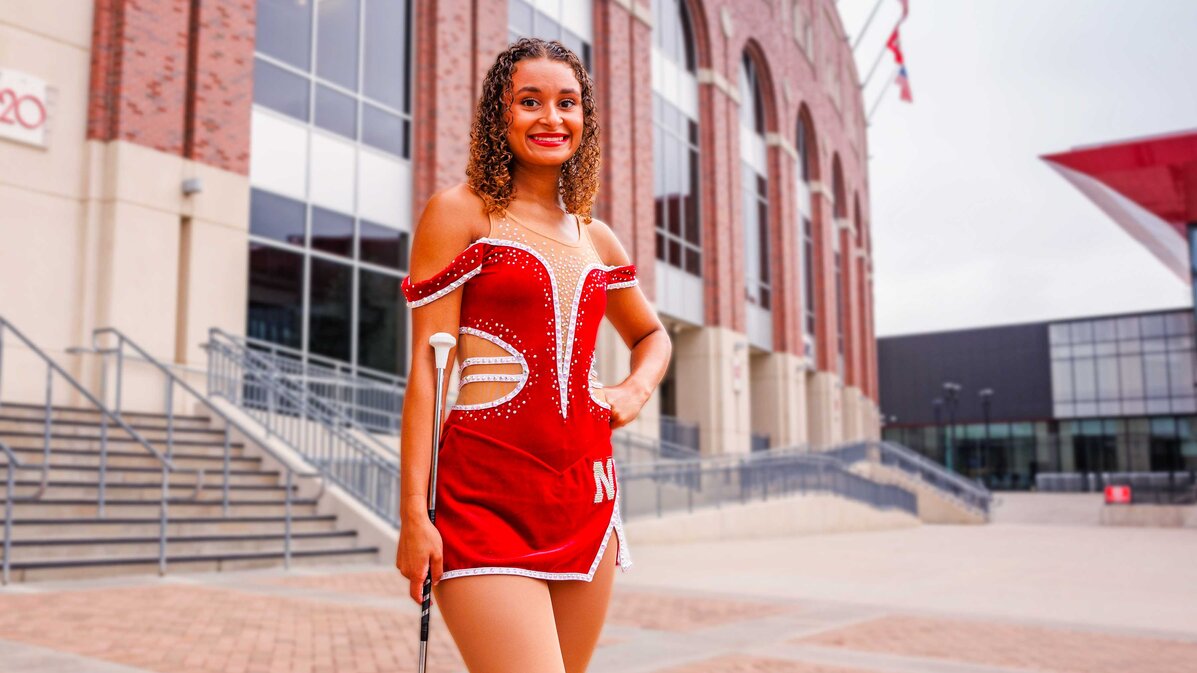 Woman in red baton twirler uniform standing outside near a brick building with stairs and flags.