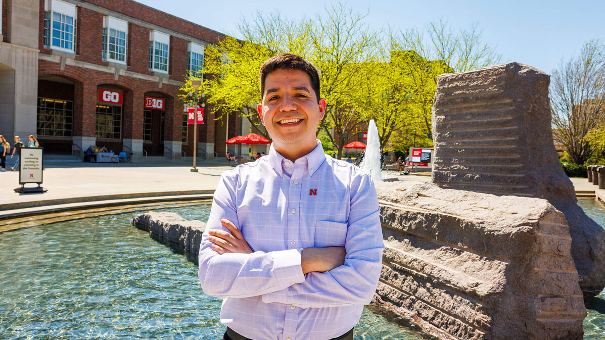 Lupe Perez standing by fountain, arms crossed on sunny day with the Nebraska Union set in the background.