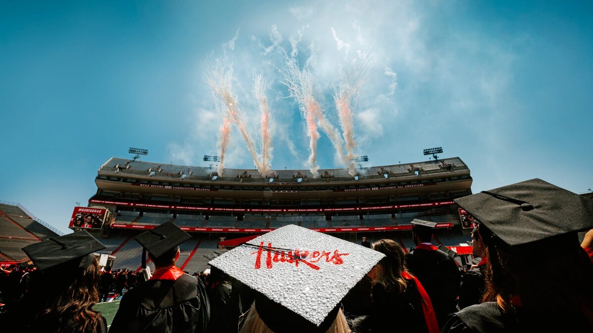 Fireworks in the shape of Ns erupt behind Memorial Stadium as graduates watch. A graduate whose mortarboard reads “Huskers” is centered in the foreground.