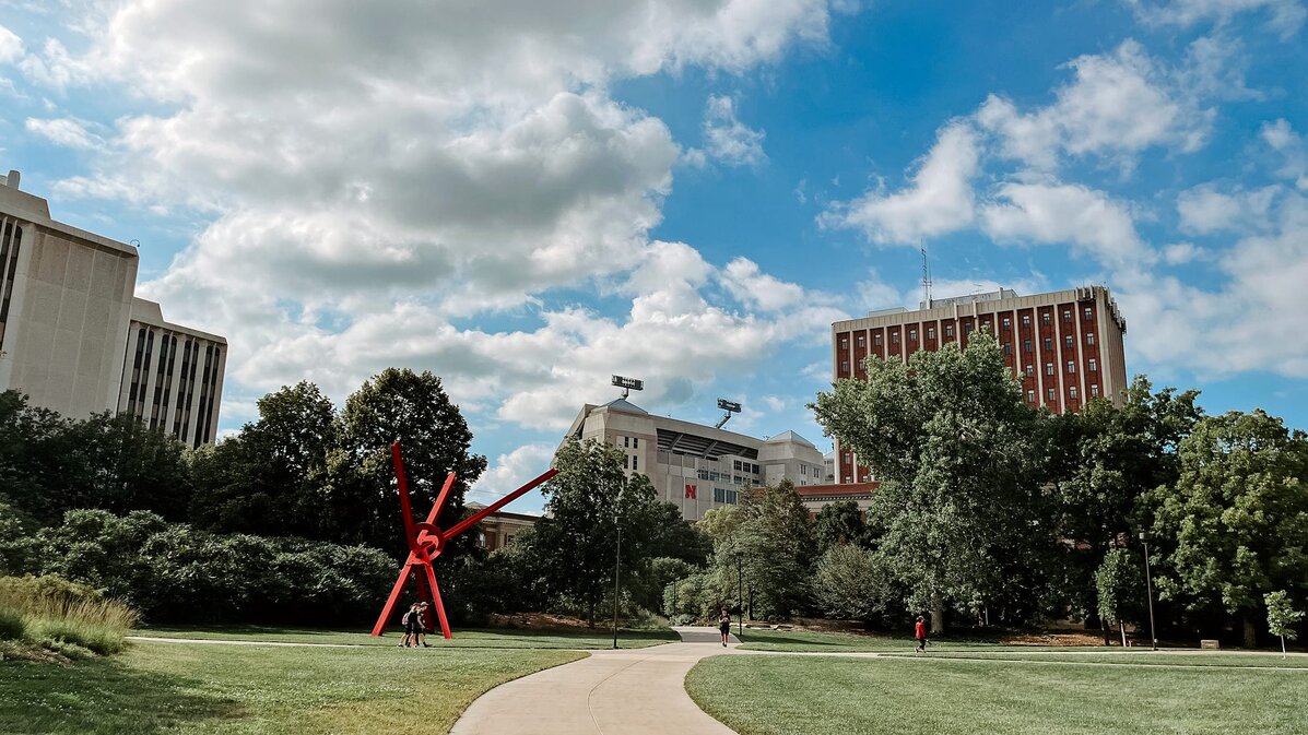 People walk along the sidewalks on campus with Memorial Stadium and Oldfather Hall in the background