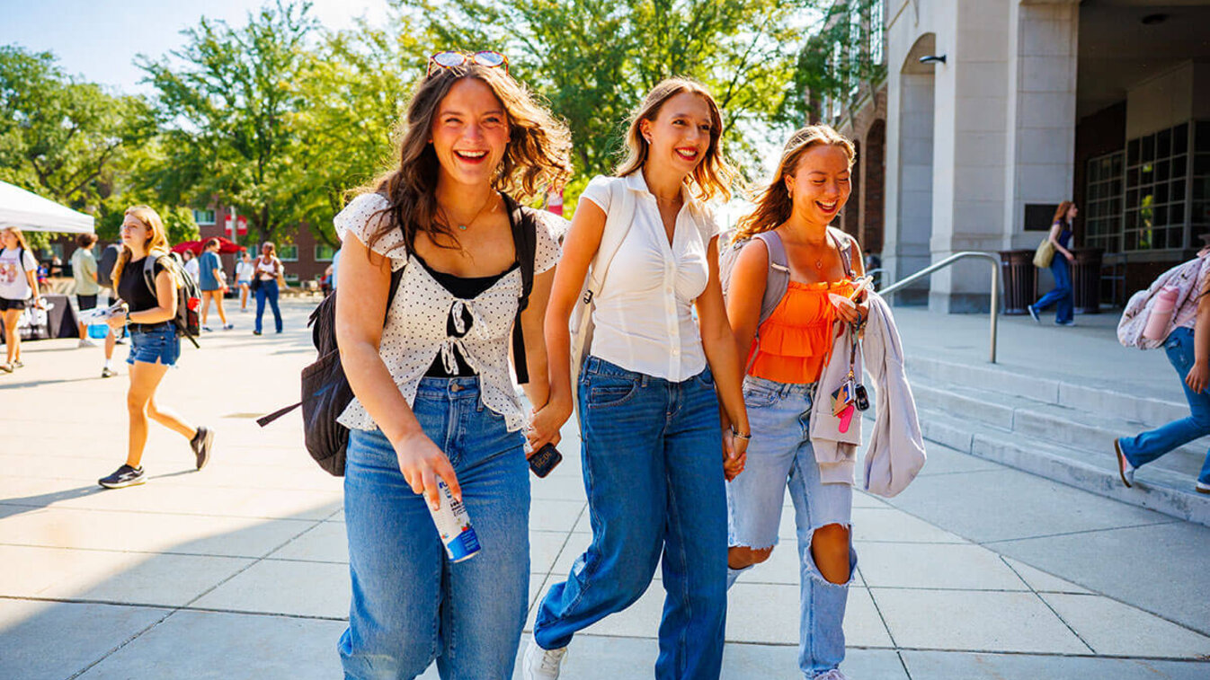 Three women smiling and walking outdoors on a sunny day, surrounded by trees and buildings.