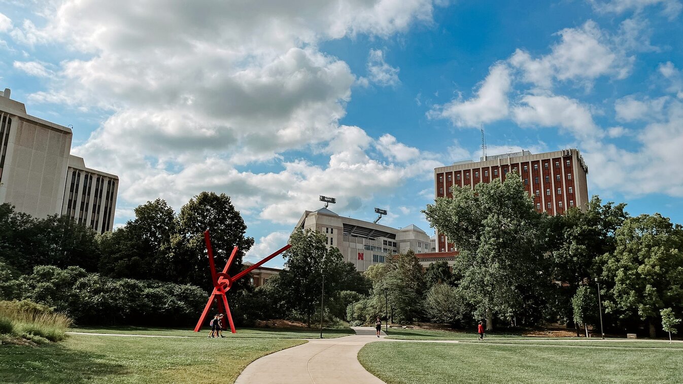 People walk along the sidewalks on campus with Memorial Stadium and Oldfather Hall in the background