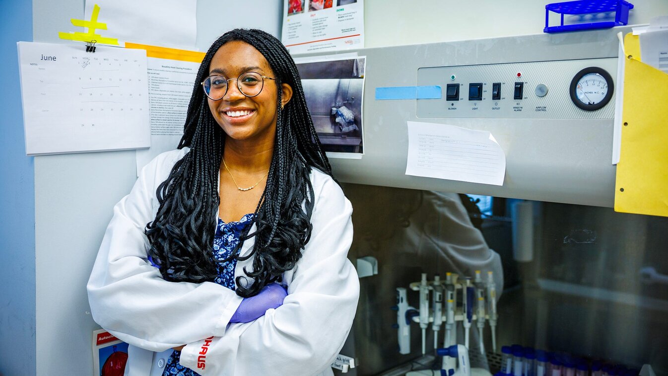 Laila smiles for a photo in a campus lab. Other photos on the page show her in the same lab, looking through a microscope and presenting a beaker to the camera