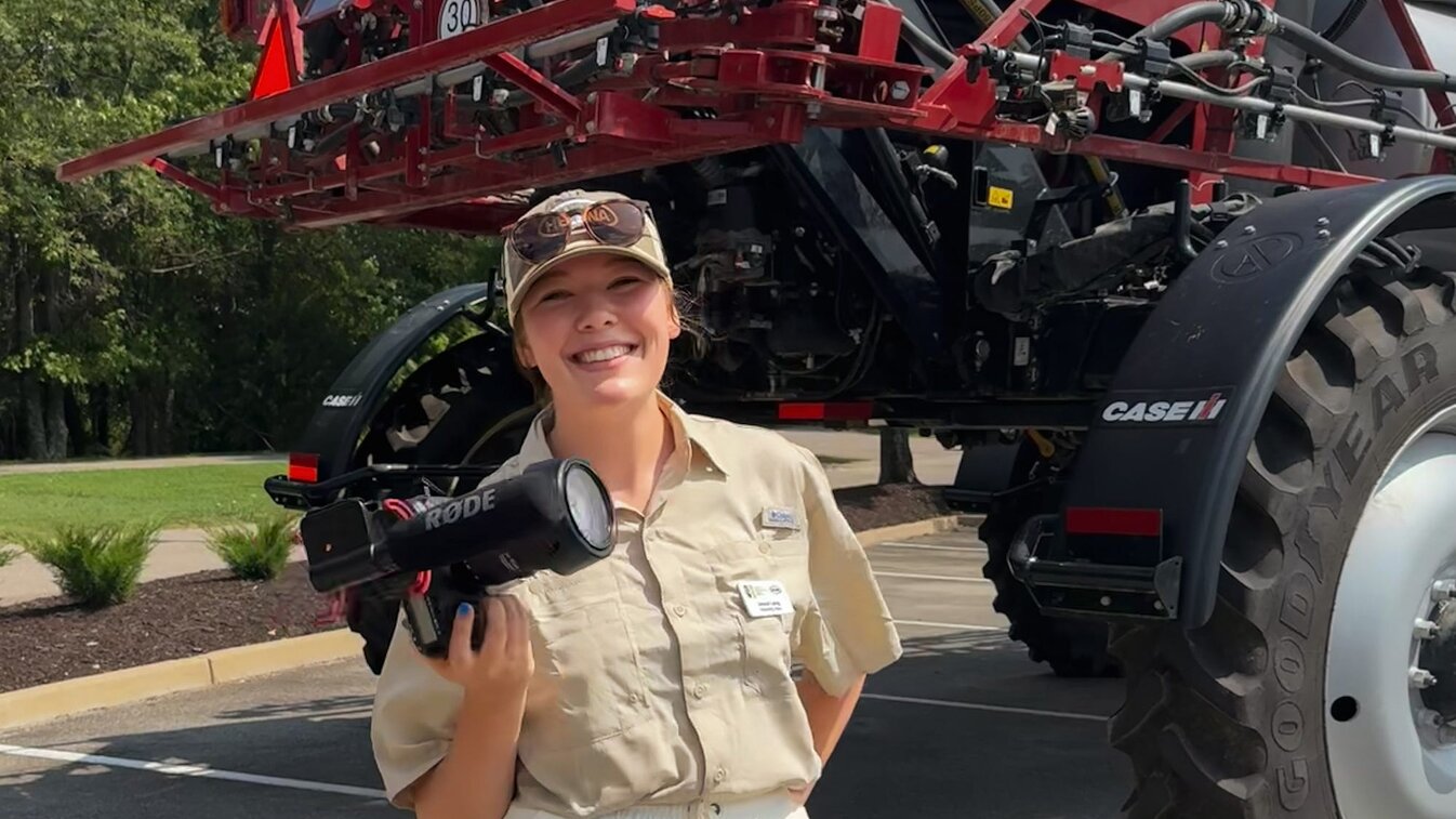 Jessie smiles for a photo holding a camera with a piece of equipment behind her