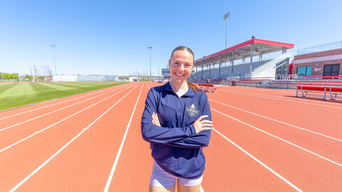 Person standing confidently on a red track field, arms crossed, with empty stadium seats in the background.