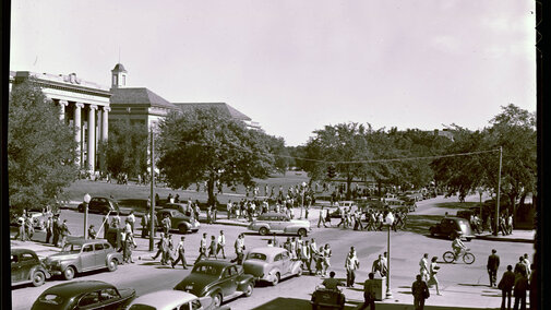 A black and white photograph of campus from the campus archives. The photo shows students walking across campus.