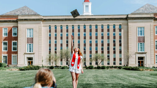 Graduate tossing cap in front of Love Library, with photographer in the foreground.