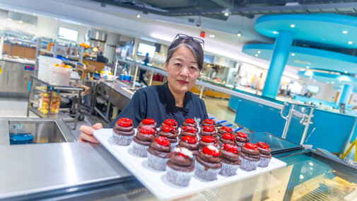 Staff member in dining hall standing in front of sheet of fancy decorated cupcakes