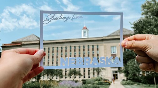 The outline of a postcard with text that says “Greetings from Nebraska” is held up to Love Library