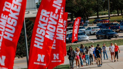 People walk along a sidewalk lined with flags that read “Husker Move-In”