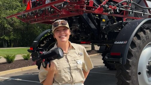 Jessie smiles for a photo holding a camera with a piece of equipment behind her