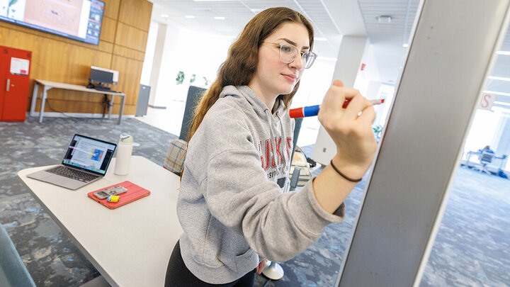 A white, brunette student with glasses, in a grey Nebraska sweatshirt, writes on a whiteboard in the library.