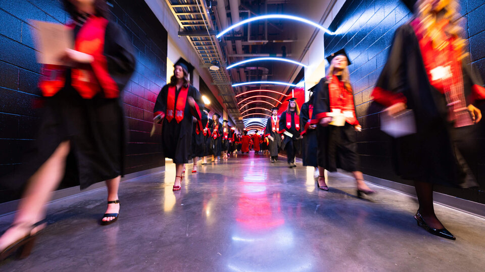 Graduates in robes and red stoles walk down a lit hallway, reflecting bright colors on the floor.