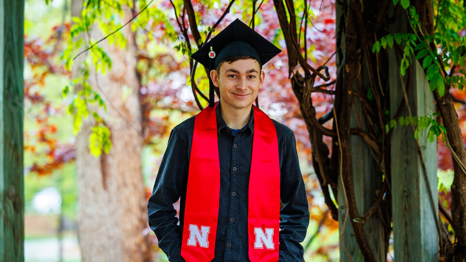 Student wearing graduation cap and sash while standing in arboretum