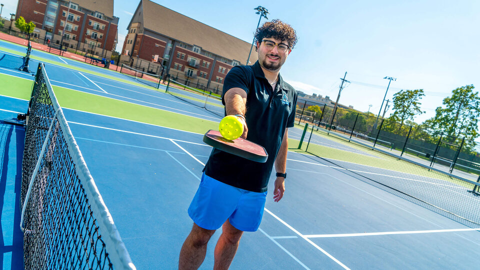 Student balancing ball on tennis racquet while standing on court in front of residence halls