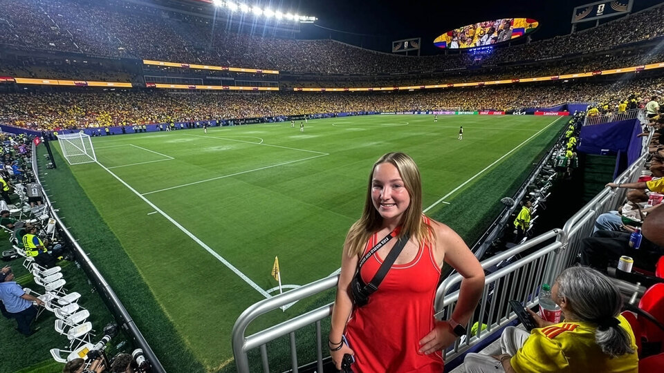 A woman in a red dress stands near a large, illuminated soccer stadium filled with fans at night.