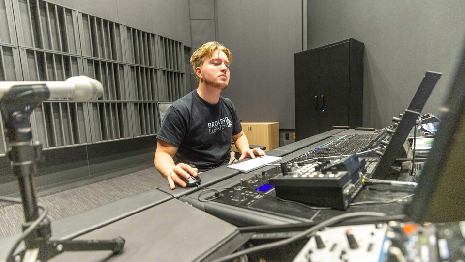 Sound engineer working at a mixing console in a studio with neutral-colored walls.