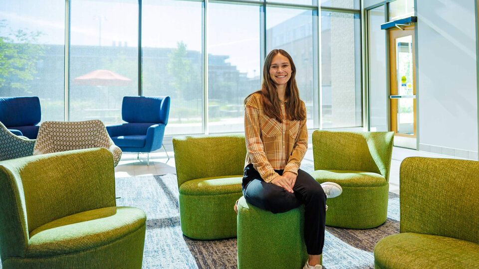 Smiling woman sitting on a green ottoman in a bright, modern lounge with large windows.