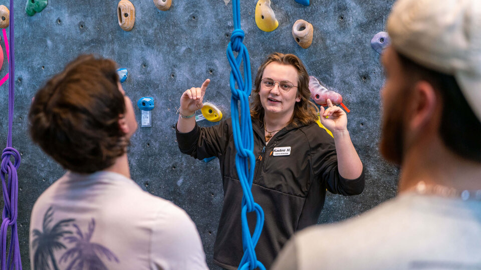 Student standing in front of climbing wall gesturing to other students