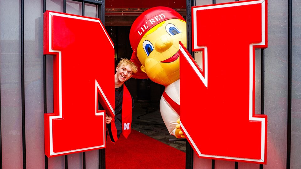 Person and Lil Red mascot peeking through large red Nebraska logo doors onto a red carpet walkway.
