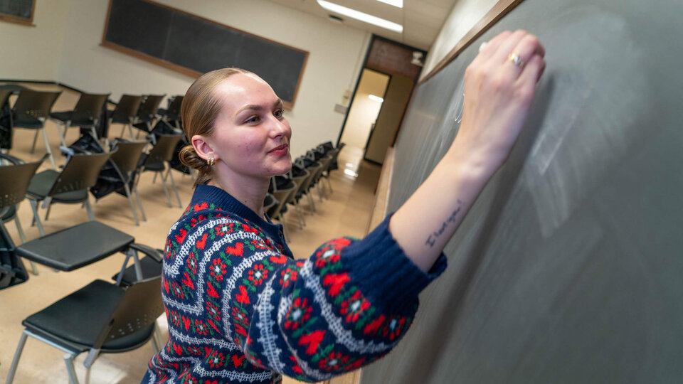 Student teacher writing on classroom chalkboard with empty chairs in background