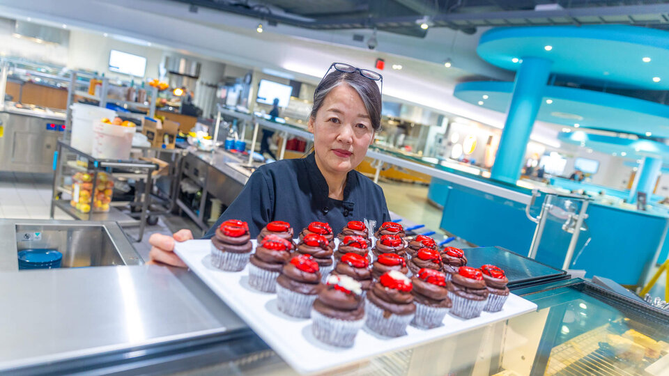 Staff member in dining hall standing in front of sheet of fancy decorated cupcakes