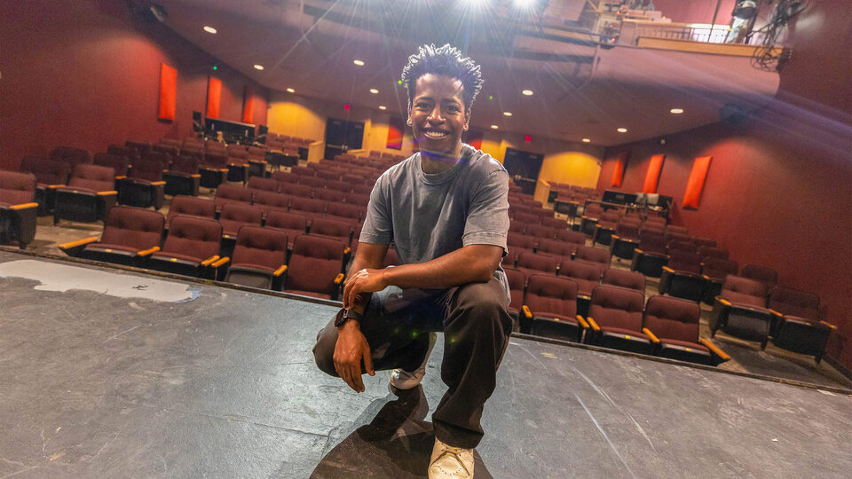 Man kneeling on a theater stage with rows of empty seats behind him.