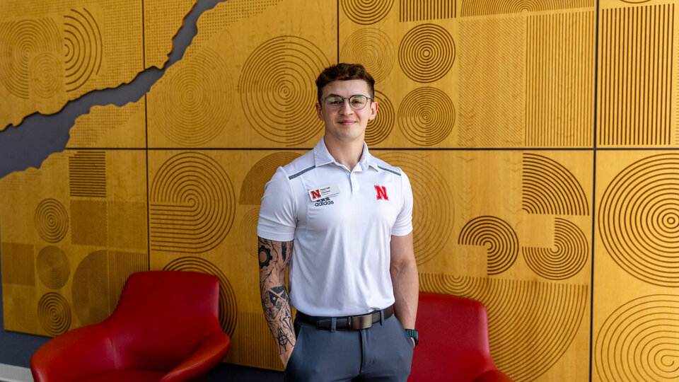 Student in white polo shirt standing in front of wooden wall