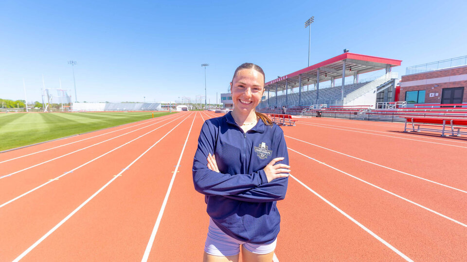 Person standing confidently on a red track field, arms crossed, with empty stadium seats in the background.