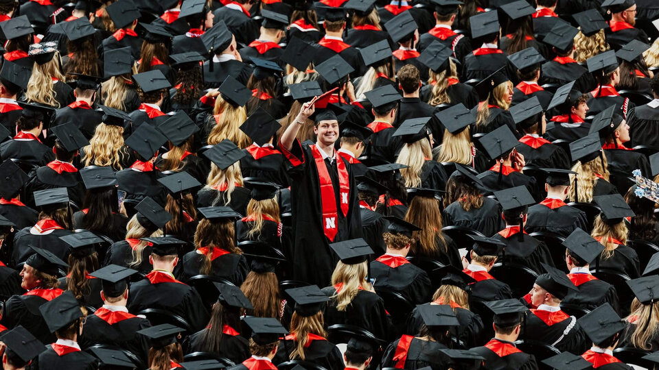 Graduates in caps and gowns, one raised hand smiling in the middle of the crowd.