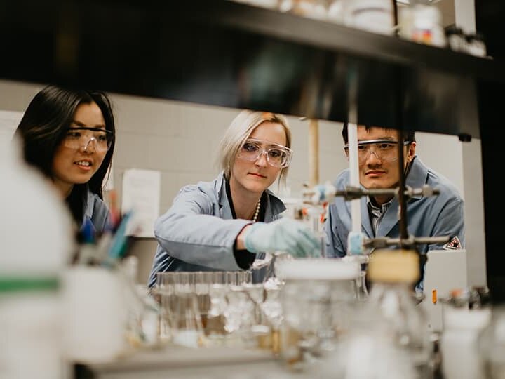 Three Huskers wearing safety goggles and light blue lab coats work in a lab
