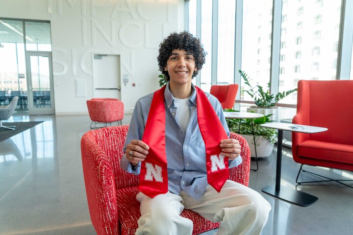 Man seated in a modern lounge holding a red Nebraska sash, with bright red chairs and large windows.