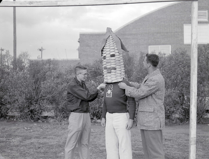 A black and white photograph of an old iteration of the Cornhusker mascot. Two individuals stand at either side as they adjust the suit of a man wearing a corncob head piece.