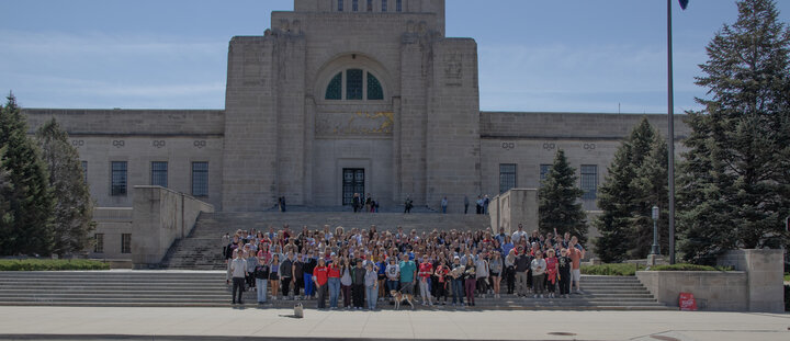 Large group gathered on stairs in front of a tall, beige building under a clear blue sky.