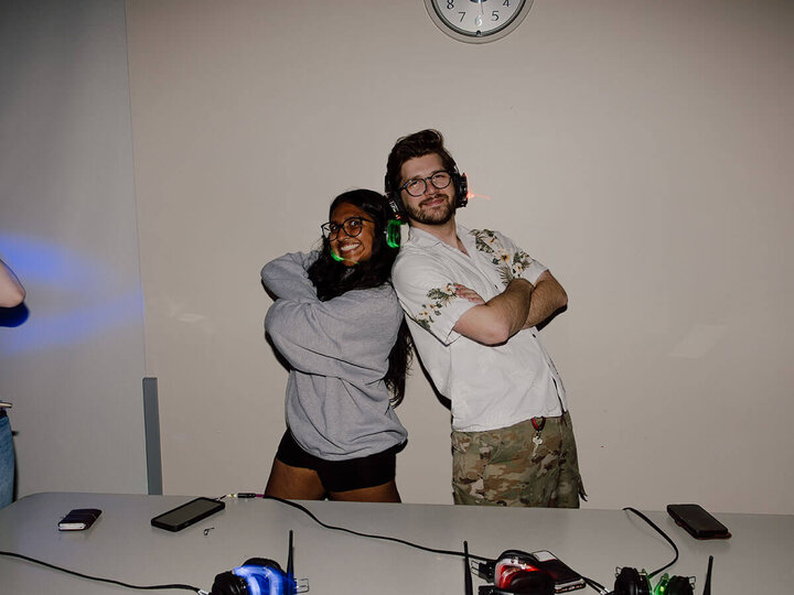 Two people smiling and posing back-to-back with crossed arms, wearing headphones in a dimly lit room.