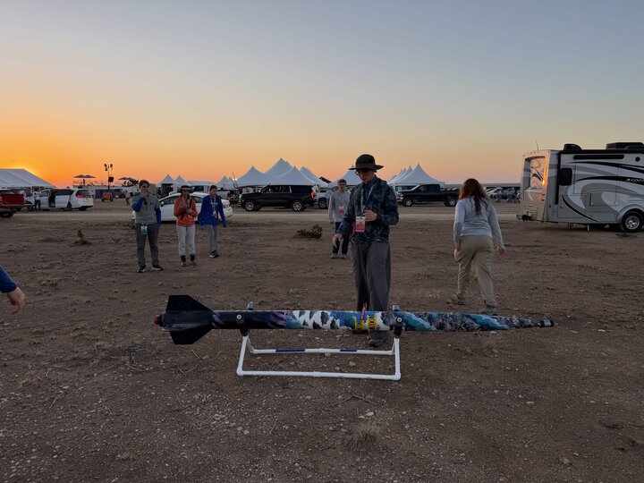 Man standing near a colorful model rocket on a stand, with a sunset and tents in the background.