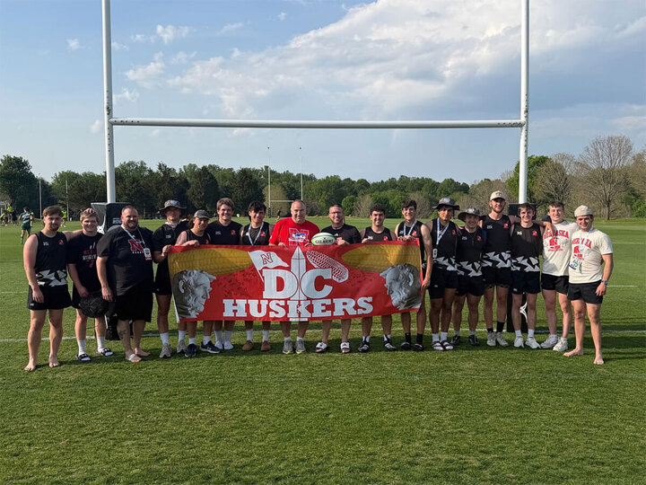Group of people holding a “DC Huskers” banner on a grassy field under a goalpost, sunny day.