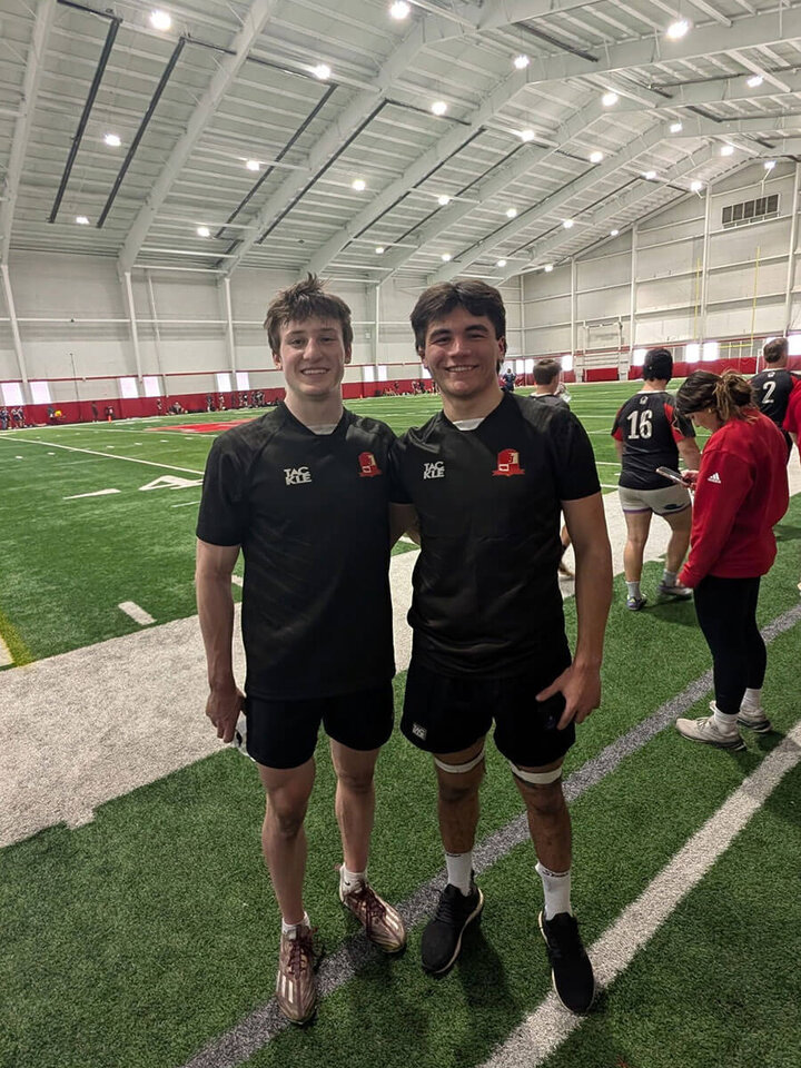 Two young men in rugby uniforms standing on an indoor field, smiling at the camera.