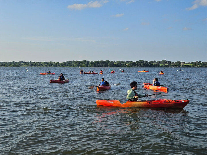 Kayakers in orange boats on a calm lake under a blue sky.