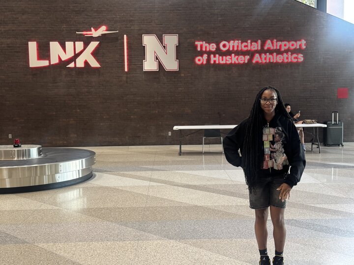 Woman standing near baggage claim in an airport with "The Official Airport of Husker Athletics" sign on wall.