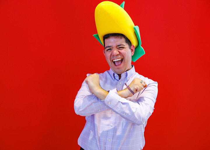 Lupe Perez in a light-colored shirt with arms crossed, wearing an inflatable corn-shaped hat against a red background.