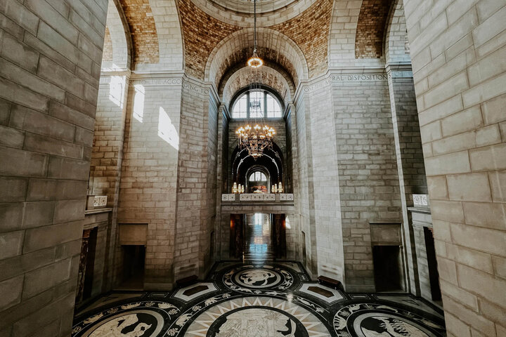 Grand hall of Nebraska State Capitol building with arched ceilings, intricate mosaic floor, and a glowing chandelier.