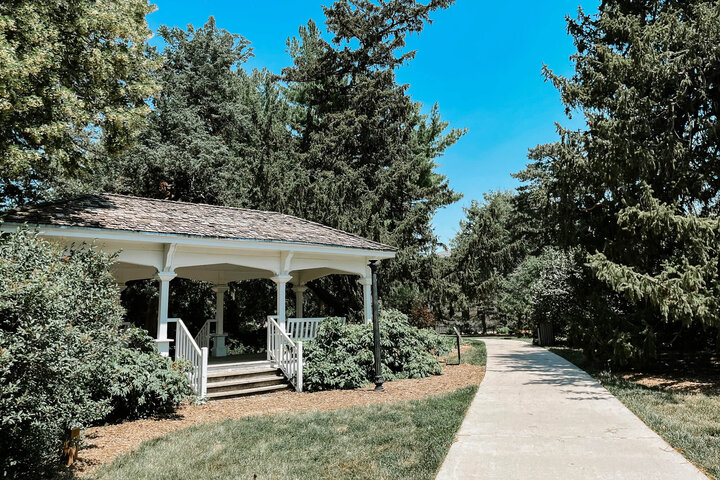 Gazebo surrounded by trees, next to a paved walkway under a bright blue sky.