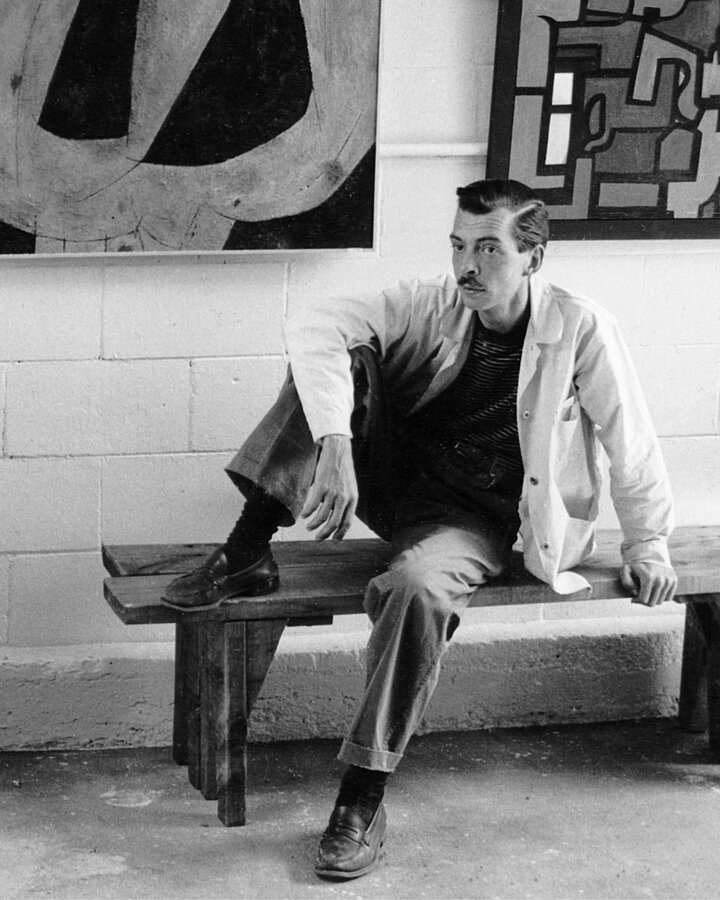 Artsist Weldon Kees sits on a bench in front of two of his modernist paintings