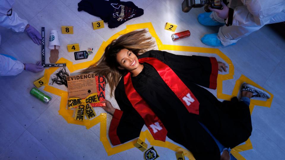 Student in graduation robe laying on ground surrounded by crime scene props