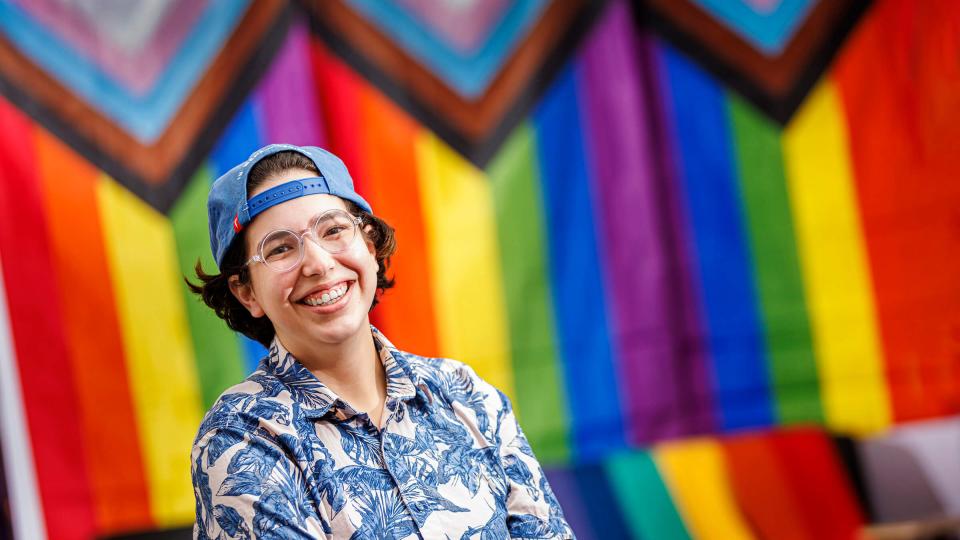 Student wearing backwards cap and blue and white shirt in front of rainbow flag
