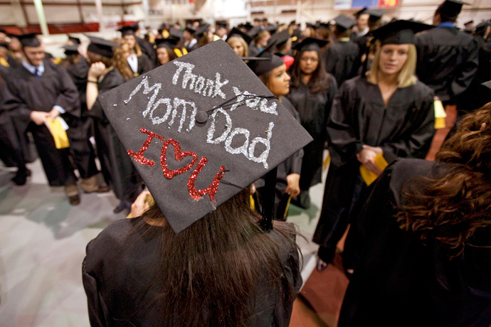 Room full of graduating students in academic regalia; focused on a decorated mortar board with the message "Thank you Mom Dad I♥U